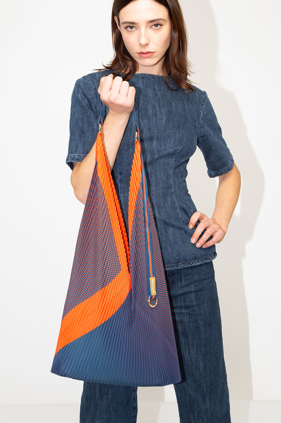 Bolso plisado Isabella Blue de Multitudes en tonos azul y naranja, con asas de satén resistentes y anilla dorada para colgar accesorios. Diseño elegante y sostenible en poliéster reciclado.