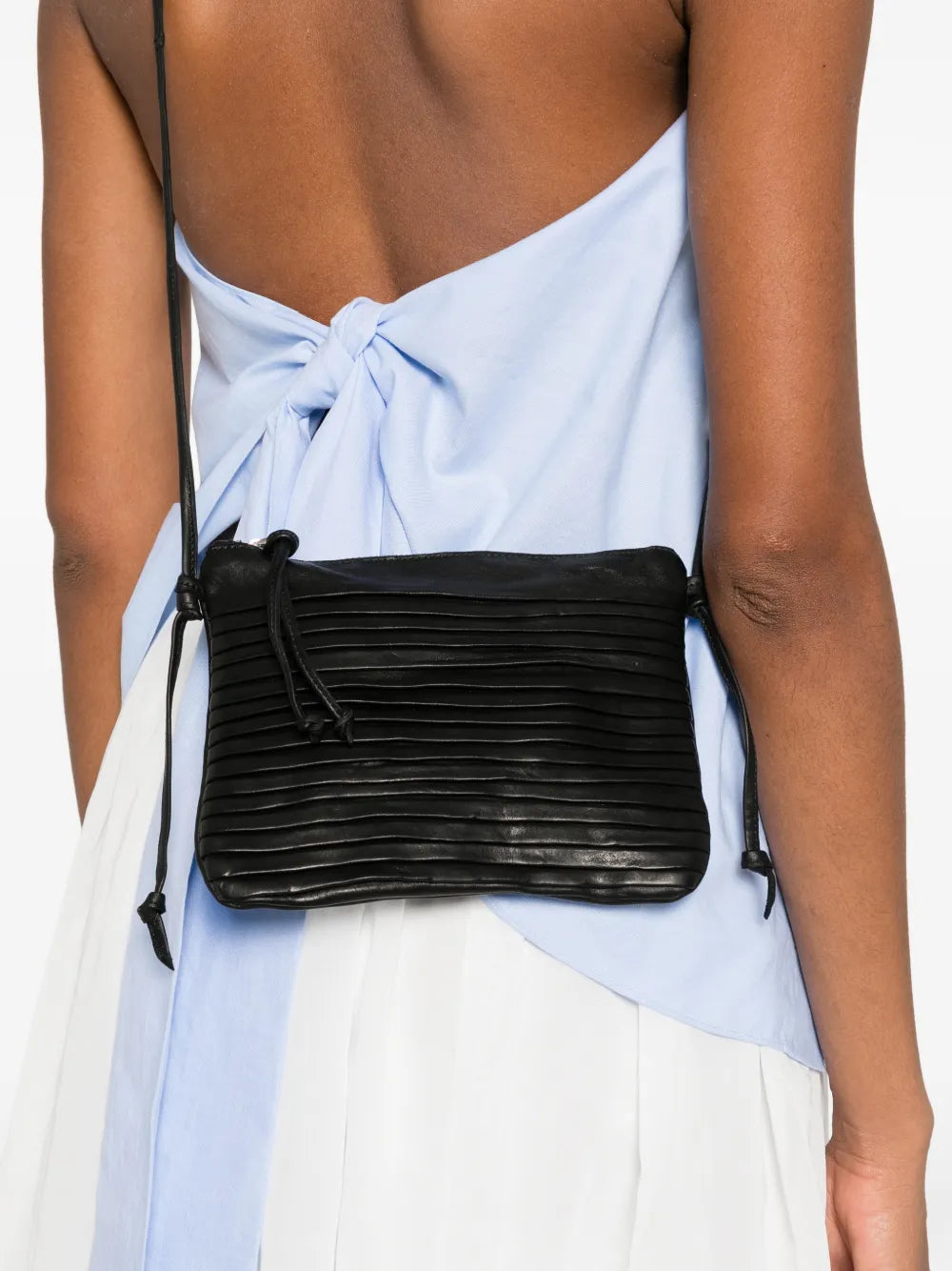 Small black leather crossbody bag with fine horizontal pleated details and adjustable strap, handcrafted in Italy, displayed against a white background.